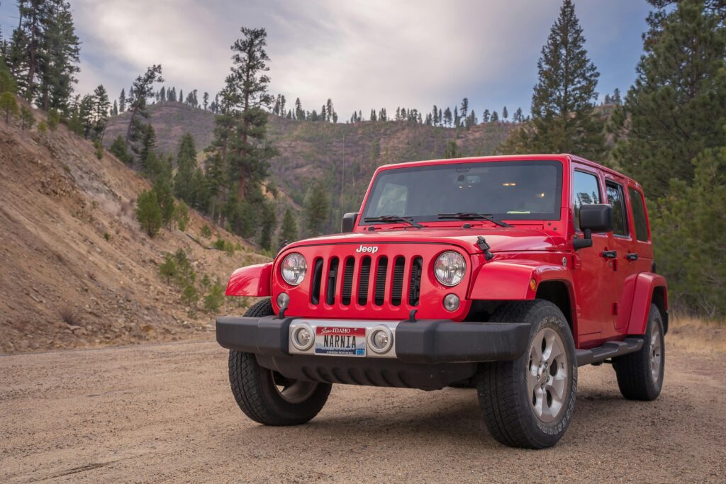 pexels-photo-1682666-1682666 Red Jeep parked on dirt road with mountainous forest backdrop, ideal for travel and adventure themes.