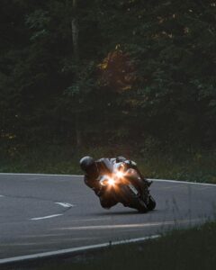 Motorcyclist leaning into a curve on a quiet road surrounded by trees, headlights on.